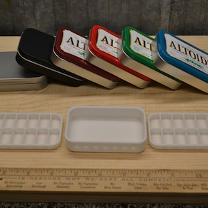 May include: Six empty Altoids tins in various colors, including silver, black, red, green, and blue, are lined up on a wooden surface.  The tins are all closed and have the Altoids logo on them.  Three white plastic containers with multiple compartments are also on the surface.