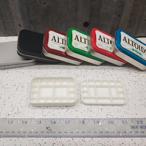 May include: Five empty Altoids tins, one silver, one black, one green, one red, and one blue, are lined up on a white surface. Two white plastic trays with 16 compartments each are in the foreground. A ruler is in the bottom of the image.