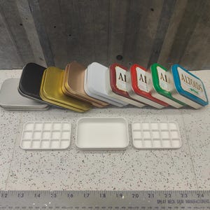 May include: A collection of Altoids tins in various colors, including silver, black, gold, and white, with the Altoids logo visible. Below are three white plastic paint palettes with multiple compartments. A ruler is at the bottom.