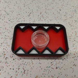 May include: A red and black tray with a clear, round container in the center. The tray has a geometric design with white triangles and black edges. The tray is on a speckled surface.