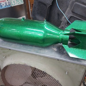 May include: A large, metallic green bomb with a glittery finish. The bomb has a cylindrical body and a finned tail section. It rests on a metal surface, with a fan-like structure visible below.