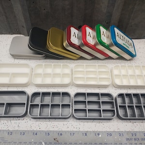 May include: A set of eight small, rectangular containers. Four are white with nine compartments each, and four are gray with nine compartments each. The containers are arranged in two rows of four. Above the containers are seven empty Altoids tins in various colors, including silver, black, gold, red, green, and blue. The blue tin has the word "Altoids" printed on it.