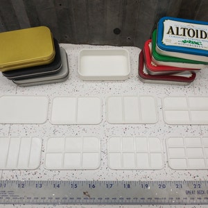 May include: Nine white plastic paint palettes of various sizes, including one with a single well, one with two wells, one with three wells, one with four wells, one with six wells, one with nine wells, and three with twelve wells. The palettes are arranged in three rows on a white surface. A ruler is visible at the bottom of the image.