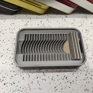 May include: A gray plastic storage case with a series of slots for holding razor blades. The case has a textured surface and a slightly curved design.