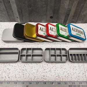 May include: Six empty Altoids tins in various colors, including red, green, blue, gold, and black, are stacked on top of each other.  Below the tins are four gray rectangular containers with different numbers of compartments.
