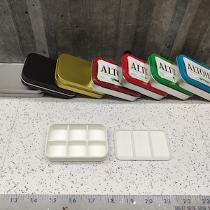 May include: A set of seven empty Altoids tins in various colors, including silver, black, gold, green, red, and blue. The tins are arranged in a row, with the largest tin at the back and the smallest tin at the front. The tins are all labeled with the word "Altoids" in white letters.