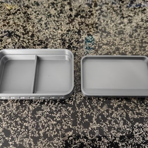 May include: A gray, rectangular container with a lid. The container is divided into two compartments and has a decorative pattern along the edge. The lid is a simple, flat rectangle. The items are made of plastic.