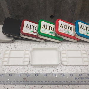 May include: Five empty Altoids tins, two silver, one black, and two with the original Altoids labels in red, green, and blue.  The tins are stacked on top of each other.  Two white plastic paint palettes with 10 wells each are in the foreground.