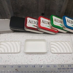 May include: Four Altoids tins in different colors: silver, black, red, green, and blue. The tins are arranged in a row, with the silver tin on the left and the blue tin on the right. The tins are all closed and have the Altoids logo on the front. There are three white rectangular trays with curved indentations on the surface.