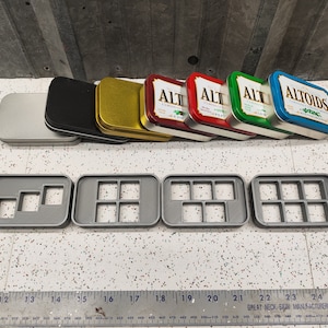 May include: Four gray plastic trays with square compartments, each tray has a different number of compartments.  The trays are on a white tiled surface.  Behind the trays are six empty metal tins, two silver, one black, one gold, and two red with the word "ALT" printed on them.  One blue tin has the word "ALTOIDS" printed on it.
