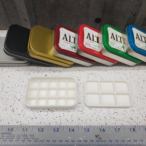 May include: Six empty Altoids tins in various colors, including silver, black, gold, red, green, and blue, stacked on top of each other. Two white plastic paint palettes with 12 and 6 wells are open and placed on a white tiled surface.