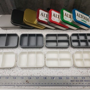 May include: Six empty metal tins with various colors, including silver, black, gold, red, green, and blue. The tins are arranged in a row. Below the tins are six empty plastic containers with three compartments each. The containers are arranged in two rows of three. A ruler is visible at the bottom of the image.