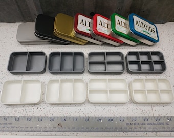 Op de afbeelding: Zes lege metalen blikken in verschillende kleuren, waaronder zilver, zwart, goud, rood, groen en blauw. De blikken zijn in een rij gerangschikt. Onder de blikken bevinden zich zes lege plastic bakjes met elk drie compartimenten. De bakjes zijn in twee rijen van drie gerangschikt. Een liniaal is zichtbaar aan de onderkant van de afbeelding.