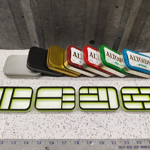 May include: Six small, rectangular metal tins with various colors and designs. The tins are arranged in a row, with the top row slightly overlapping the bottom row. The tins are all empty and have a slightly worn look. The tins are labeled with the word "Altoids" in white letters on a blue background.  The tins are sitting on a white surface with a ruler in the foreground.