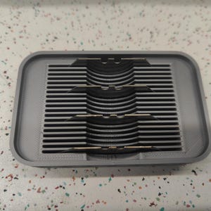 May include: A rectangular, gray 3D-printed object with a series of parallel horizontal lines and three curved, black, slotted sections. The object has a raised border and is likely a functional item.