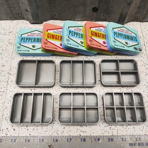 May include: Six small, round, metal tins with lids, each containing organic pastilles. The tins are arranged in a row, with the lids open. The tins are labeled with the flavors: Peppermint, Ginger, and Pepper. The tins are on a gray surface.