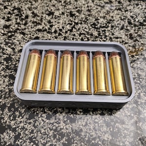 May include: A gray 3D-printed ammunition holder containing six brass-colored bullets. The bullets have a copper-colored tip and are arranged neatly in individual slots within the rectangular holder. The holder sits on a speckled surface.