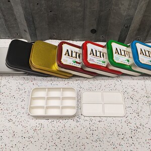 May include: A set of seven empty Altoids tins in various colors, including silver, black, gold, red, green, and blue. The tins are stacked on top of each other. The tins are labeled with the word "Altoids" and the flavor of the mints. Two white plastic trays with multiple compartments are in the foreground.