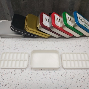 May include: Six empty metal tins stacked on top of each other. The tins are silver, black, gold, red, green, and blue. The blue tin has the word "Altoids" printed on it. Three white plastic trays with 16 compartments each are in the foreground.