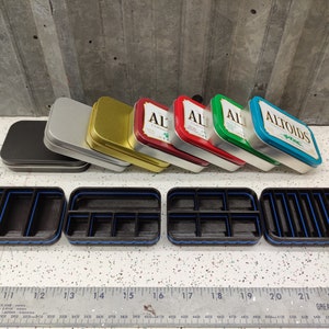 May include: Six small, rectangular metal tins with various colors, including silver, gold, green, red, and blue. The tins are stacked on top of each other. The top tin is blue and has the word "Altoids" printed on it. The tins are sitting on a white tiled surface. There are four black rectangular boxes with blue trim and multiple compartments.