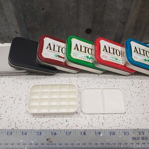 May include: Five empty Altoids tins in different colors, including red, green, blue, and silver. The tins are arranged in a row on a white surface. The tins are labeled with the text "Altoids Curiously Strong Mints" and the flavor of the mints. There are two white plastic trays with compartments, one with 16 compartments and the other with two large compartments.