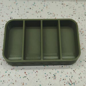 May include: An olive green rectangular organizer with four compartments. The base features a row of star-shaped cutouts. The rounded corners and vertical dividers create a structured storage solution.
