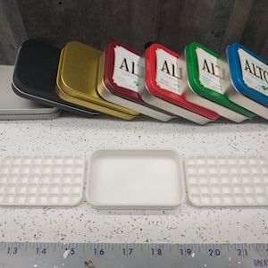 May include: Seven empty metal tins with various colors and designs, including the text "Altoids" and "Car-Freshner", are lined up on a white surface. Three white plastic trays with square compartments are in the foreground.