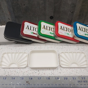 May include: Six empty Altoids tins, three stacked on top of each other, and three in a row. The tins are silver with red, green, and blue lids. The lids have the Altoids logo and flavor names:  "Curiously Strong Mints Cinnamon", "Curiously Strong Mints Spearmint", "Curiously Strong Mints Peppermint", and "Curiously Strong Mints Wintergreen". Three white rectangular trays with a sunburst design are in front of the tins.