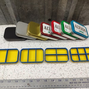 May include: Four blue and yellow plastic containers with dividers, next to seven empty metal tins. The tins are stacked and have various colors, including silver, gold, red, green, and blue. The tins have the word "Altoids" printed on them.