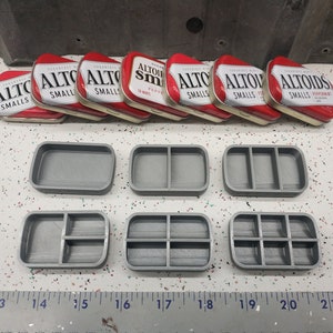 May include: Eight Altoids tins in various flavors, including peppermint, wintergreen, and cinnamon, are arranged in a row on a white surface. Below the tins are six gray 3D printed containers with different compartment configurations.