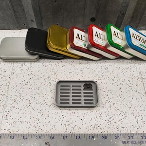 May include: A collection of six small, rectangular metal tins with various colors and designs. The tins are stacked on top of each other. The top tin is blue and says "Altoids" in white letters. The other tins are gold, red, green, and silver. The tins are on a white tiled surface. There is a gray plastic tray with a grid pattern below the tins.