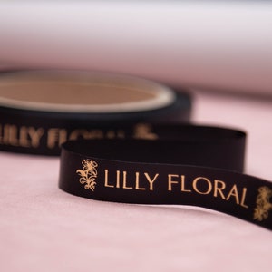 Puede incluir: Cinta de satén negra con letras doradas que dicen "Lilly Floral" y un diseño floral dorado.