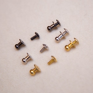 May include: Assortment of metal rivets in silver, gold, and black finishes. The rivets have a round head and a flat base.