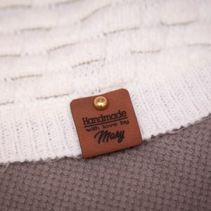 Pode incluir: Uma etiqueta de couro marrom com um rebite dourado e o texto "Handmade with love by Mary" em letras pretas.