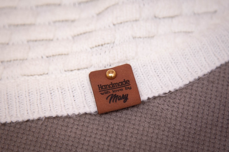 Pode incluir: Uma etiqueta de couro marrom com um rebite dourado e o texto "Handmade with love by Mary" em letras pretas.