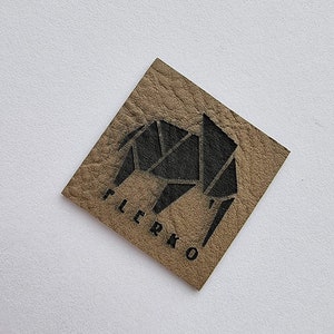 K&ouml;nnte beinhalten: Ein braunes Leder-Patch mit einem schwarzen geometrischen Elefanten-Design und dem Wort "FLERKO" in Schwarz gedruckt.