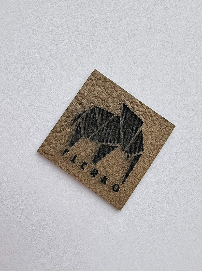 K&ouml;nnte beinhalten: Ein braunes Leder-Patch mit einem schwarzen geometrischen Elefanten-Design und dem Wort "FLERKO" in Schwarz gedruckt.