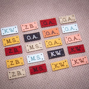 May include: Assortment of colorful leather labels with different initials, including KW, ZB, MS, and OA. The labels are arranged in rows on a textured surface.