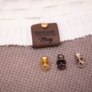 Pode incluir: Uma etiqueta de couro marrom com o texto "Handmade with love by Mary" e um rebite dourado. Tr&ecirc;s bot&otilde;es de press&atilde;o met&aacute;licos, um dourado e dois prateados, s&atilde;o mostrados em uma superf&iacute;cie marrom texturizada.