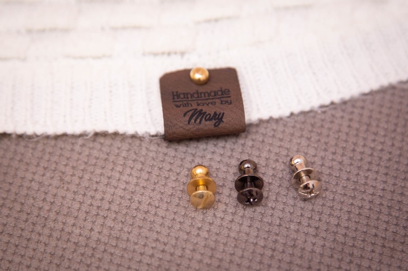 Pode incluir: Uma etiqueta de couro marrom com o texto "Handmade with love by Mary" e um rebite dourado. Tr&ecirc;s bot&otilde;es de press&atilde;o met&aacute;licos, um dourado e dois prateados, s&atilde;o mostrados em uma superf&iacute;cie marrom texturizada.