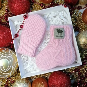 K&ouml;nnte beinhalten: Ein Paar rosa gestrickte Babyschuhe und F&auml;ustlinge mit einem kleinen schwarzen Etikett, auf dem "Woolys" auf dem Schuh steht.