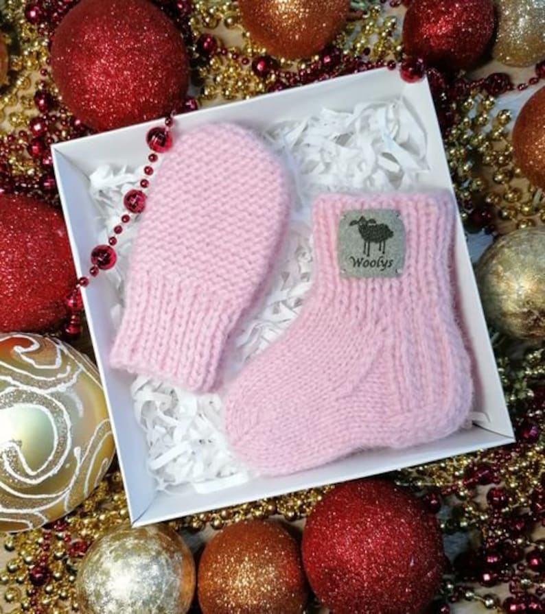 K&ouml;nnte beinhalten: Ein Paar rosa gestrickte Babyschuhe und F&auml;ustlinge mit einem kleinen schwarzen Etikett, auf dem "Woolys" auf dem Schuh steht.