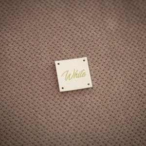 K&ouml;nnte beinhalten: Ein kleines, quadratisches, beigefarbenes Etikett mit abgerundeten Ecken und dem Wort "White" in goldenen Buchstaben.