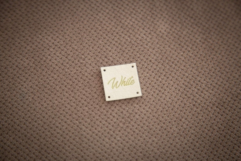 K&ouml;nnte beinhalten: Ein kleines, quadratisches, beigefarbenes Etikett mit abgerundeten Ecken und dem Wort "White" in goldenen Buchstaben.