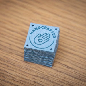 K&ouml;nnte beinhalten: Ein Stapel blauer quadratischer Etiketten mit dem Wort "HANDCRAFTED" in einem Kreis um eine Hand mit einem Herzen in der Handfl&auml;che.