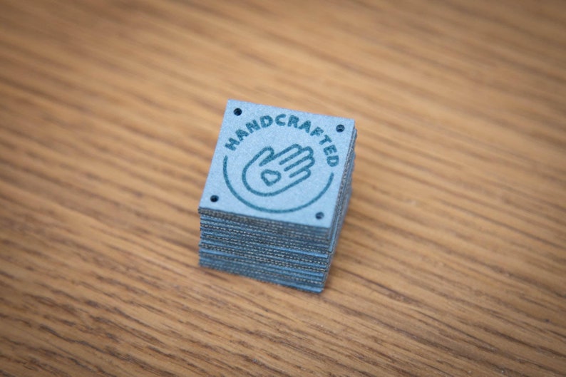 K&ouml;nnte beinhalten: Ein Stapel blauer quadratischer Etiketten mit dem Wort "HANDCRAFTED" in einem Kreis um eine Hand mit einem Herzen in der Handfl&auml;che.