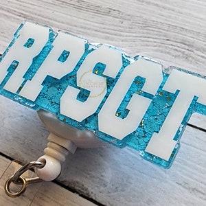 Peut inclure: Un enrouleur de badge avec un embout rectangulaire en résine turquoise pailletée portant les lettres blanches RPSGT en relief. Le mécanisme de l'enrouleur est en plastique blanc avec une pince métallique argentée fixée à la base.