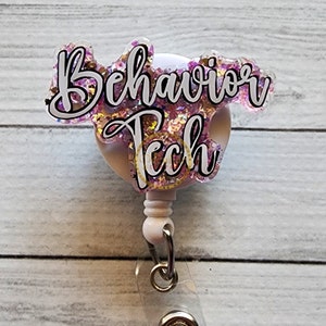 Puede incluir: Un portatarjetas con las palabras "Behavior Tech" en una fuente cursiva. El texto está delineado en negro y relleno de purpurina rosa. El carrete está sujeto a un clip de plástico transparente.