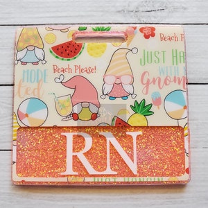 May include: A pink and white glitter badge holder with a summer gnome theme. The badge holder has a clear window for displaying a name tag. The text on the badge holder reads "Beach Please!" and "Just hang with a gnome".