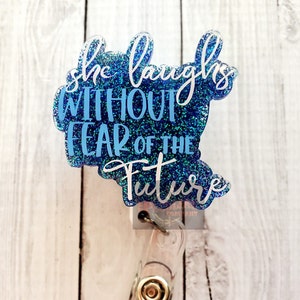 Puede incluir: Un portatarjetas azul brillante con la cita "she laughs WITHOUT FEAR OF THE Future" en letras blancas y azules. El portatarjetas tiene un cordón retráctil de plástico transparente y un clip metálico.